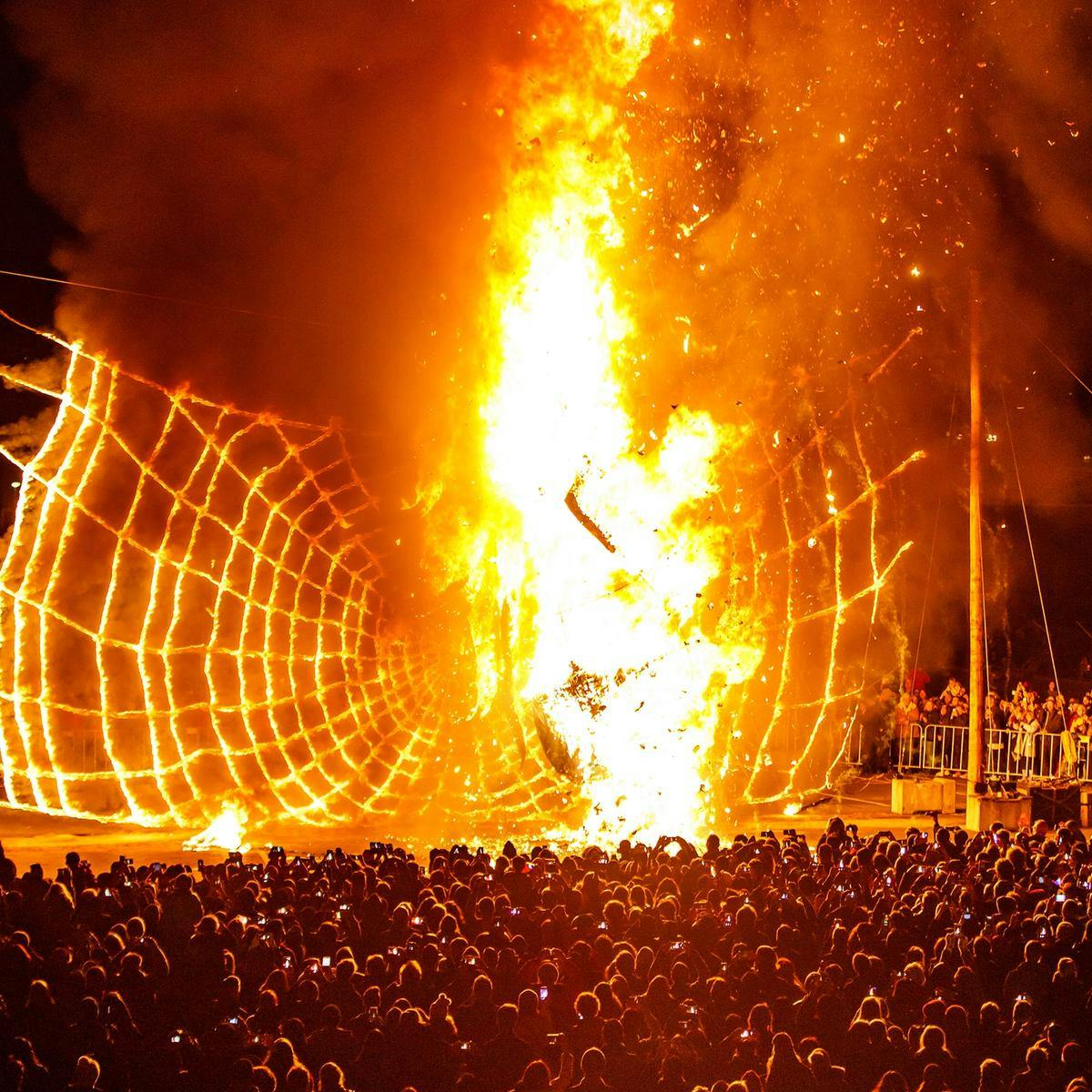 A large crowd of thousands of people watch as a spider Ogoh-Ogoh sculpture is engulfed in bright orange flames.