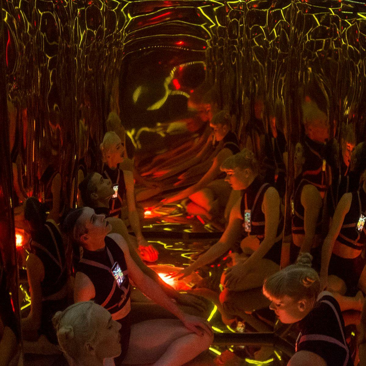 Two people wearing phones strapped to their chest sit on the floor of a small golden mirrored room.