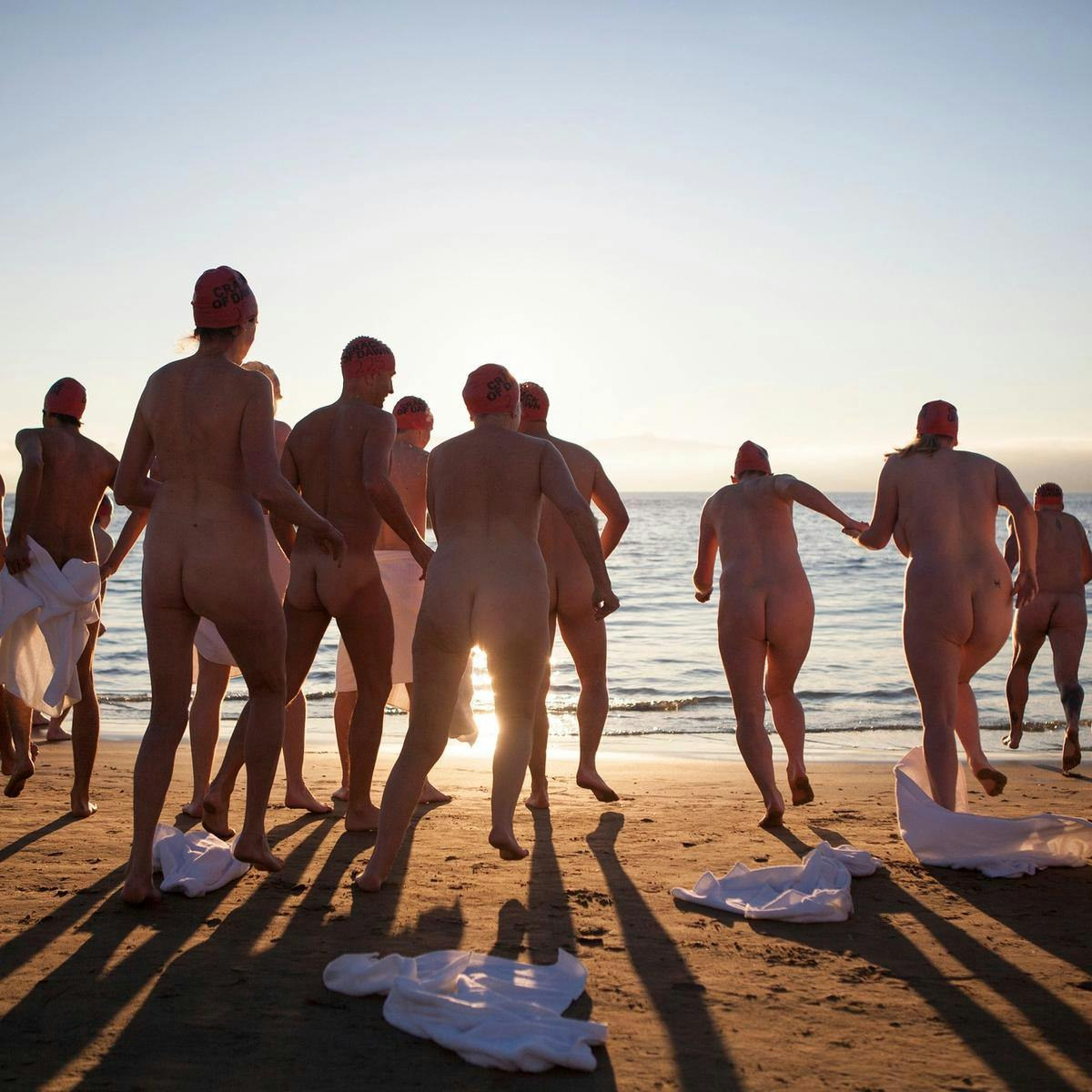 Several naked people drop their towels as they run into the ocean.