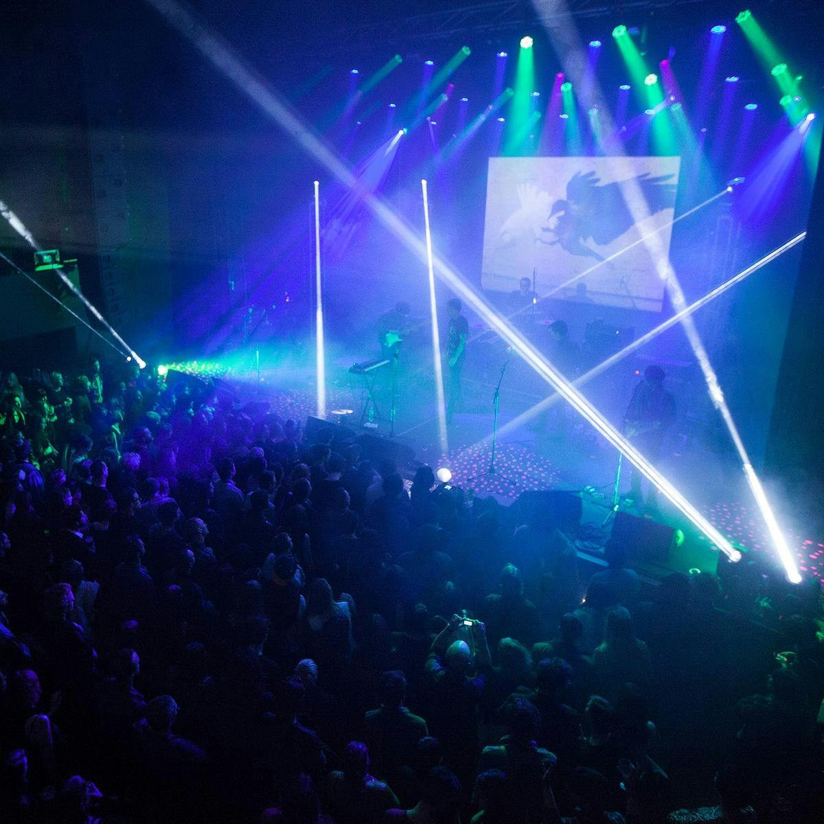 A large crowd of hundreds watch several music performers on stage. White and green beams light up the musicians. 