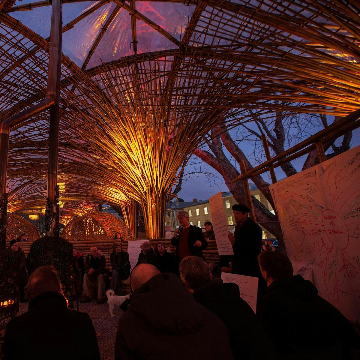 Several people sit listening to a public speaker, within an ornate bamboo structure. A detailed mind-map is drawn up on a large sheet of paper.