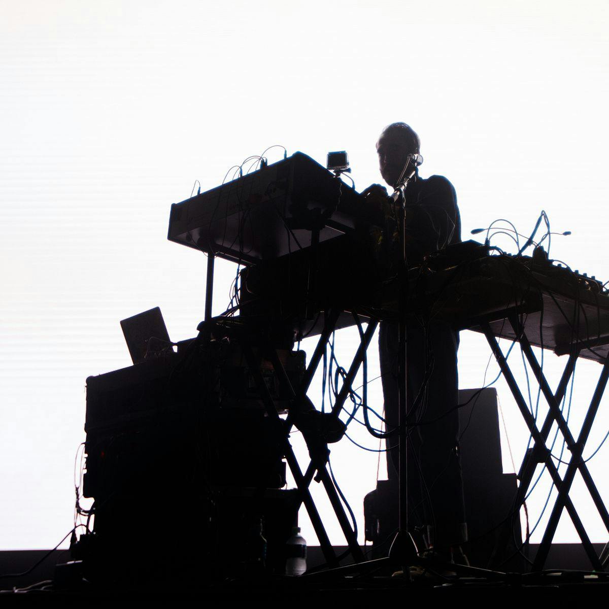 A silhouetted figure on stage stands behind a music workstation, cables can be seen connecting and running between several devices.