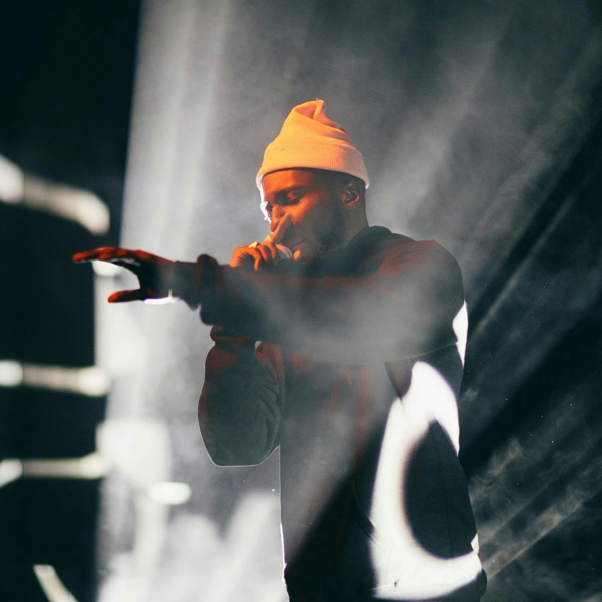 A person wearing a beanie performs into a microphone, various shapes are projected, hitting them and the background behind.