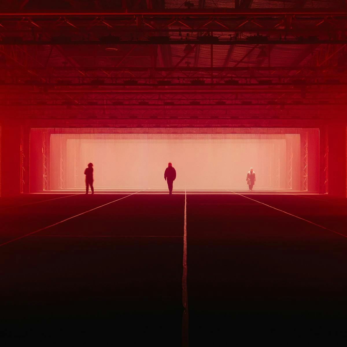 Three people walk along a wide warehouse, red light fills out the space behind them.