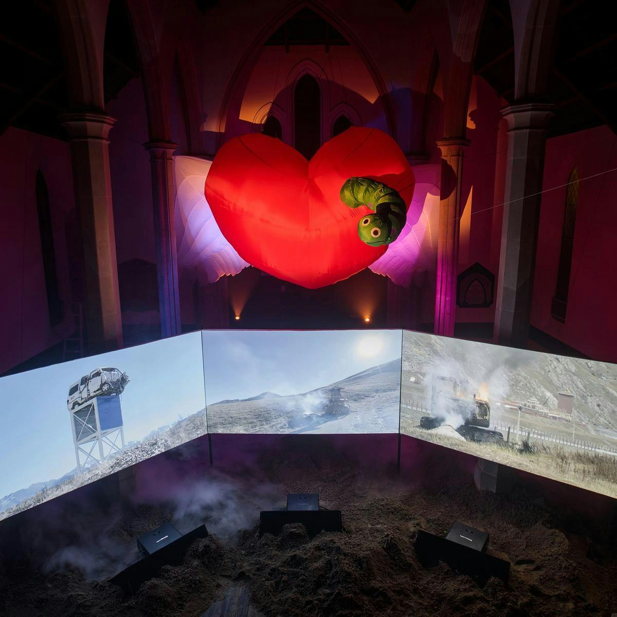 Hanging from the ceiling of a church, a large winged heart has a green cartoon worm protruding from it. At ground level three large screen show various damaged machinery.