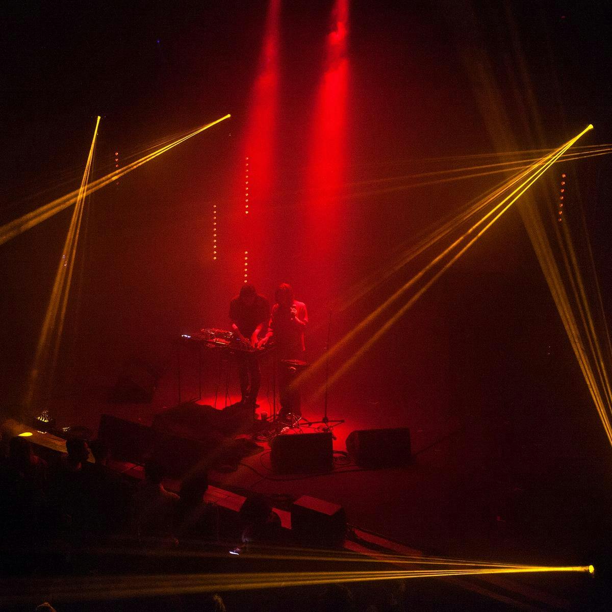 Two people perform on-stage in front of a large crowd, two red spotlights shine down on them. Several other orange lights project from above the stage.