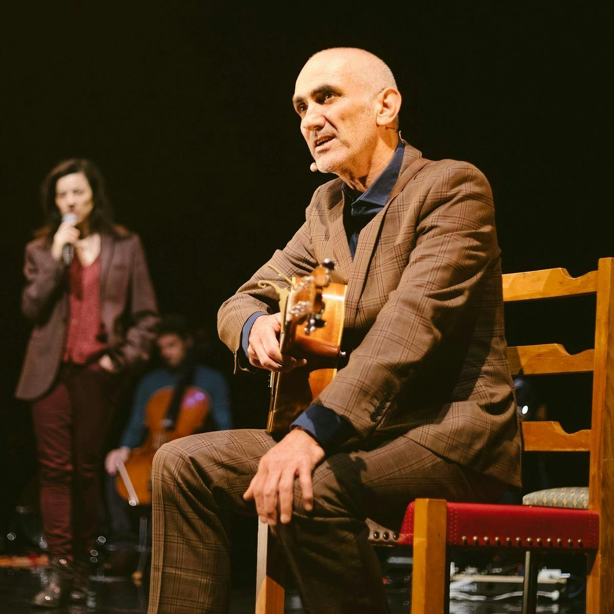 Paul Kelly sits on a wooden chair, guitar resting on his lap.