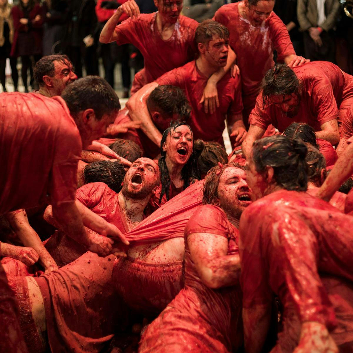 Several people covered in blood writhe on the ground, clambering on each-other, their clothes completely soaked red.