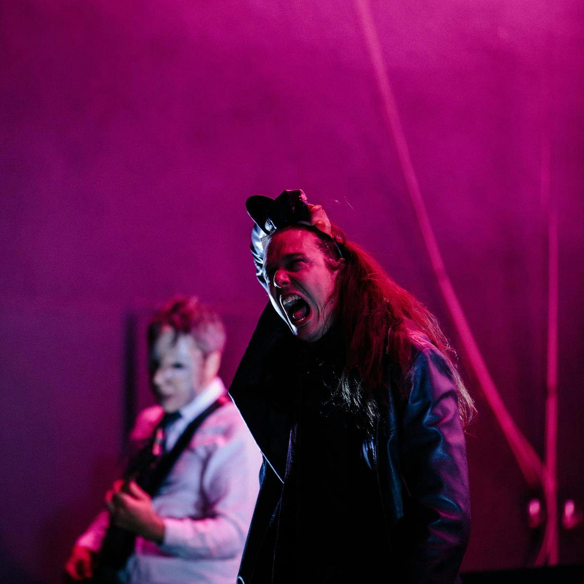 A person wearing a long leather jacket screams, in the background a masked figure plays guitar.