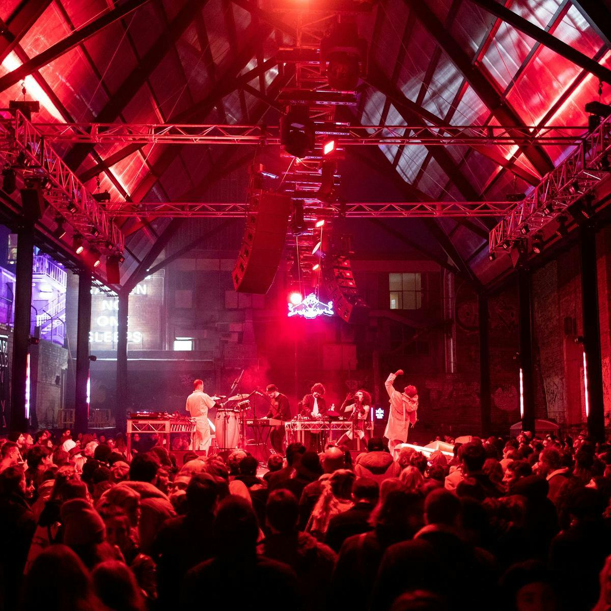 Several people perform from a raised stage for a large crowd. An a-frame roofed structure reaches high above them, from where speakers and red lights hang from.