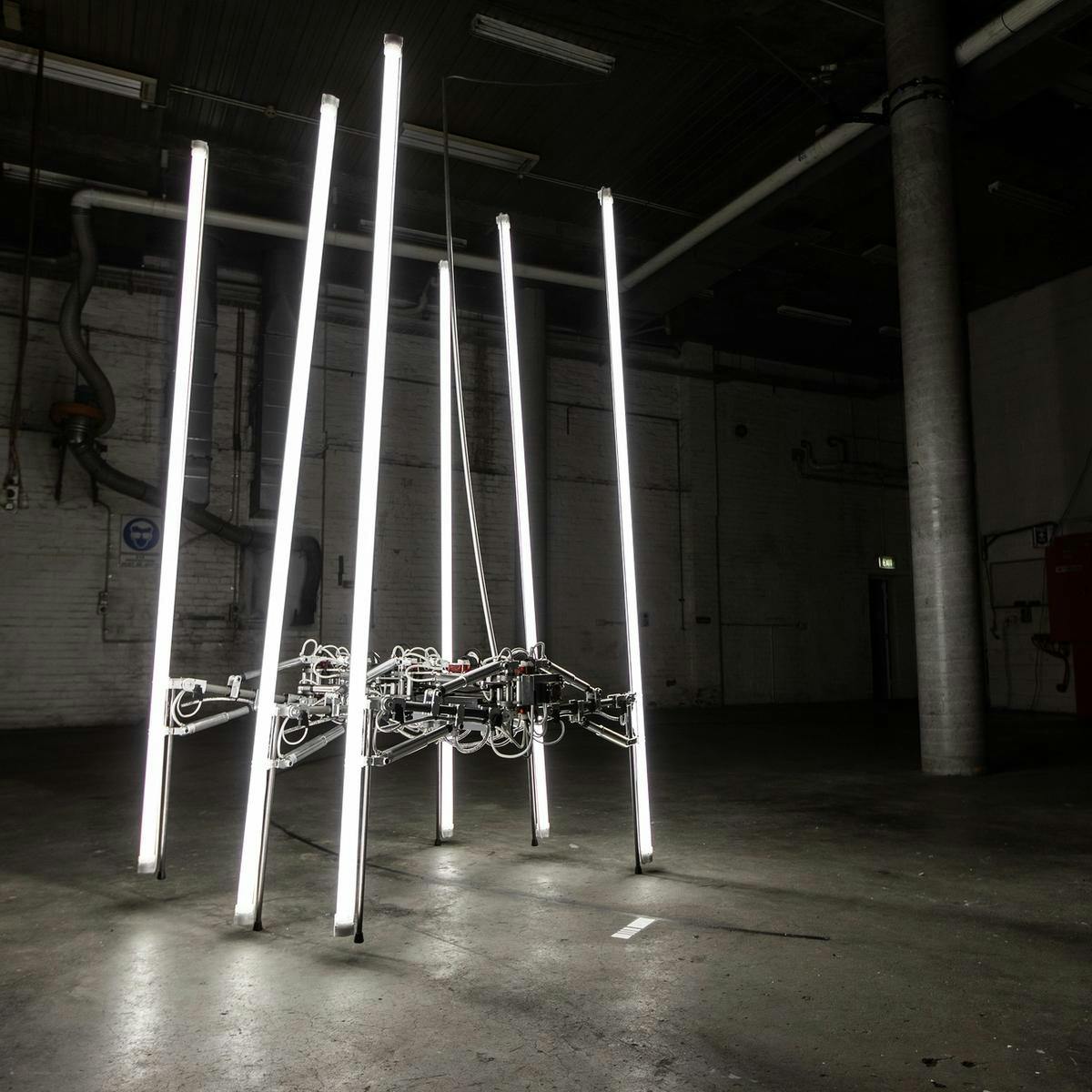 A futuristic spider-like object made up of metal and wires, stands alone is a room. Its six legs are fluorescent light tubes, providing a light source.