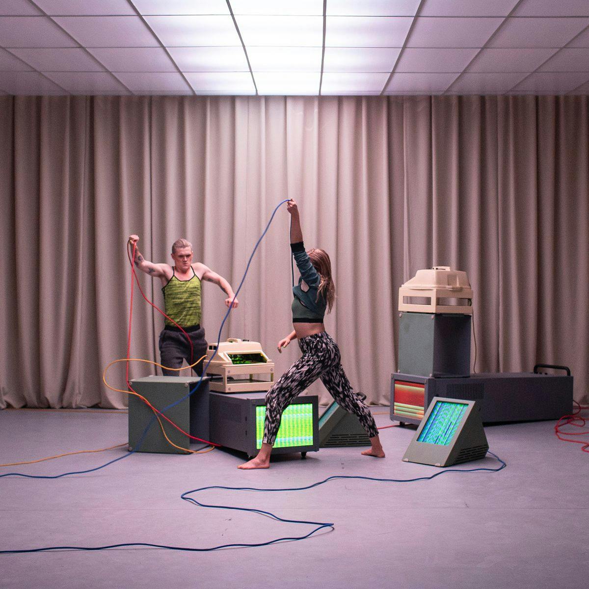 Two performers act out a scene in a small room. Several screens and wires are strewn around the floor, with brown curtains lining the walls.