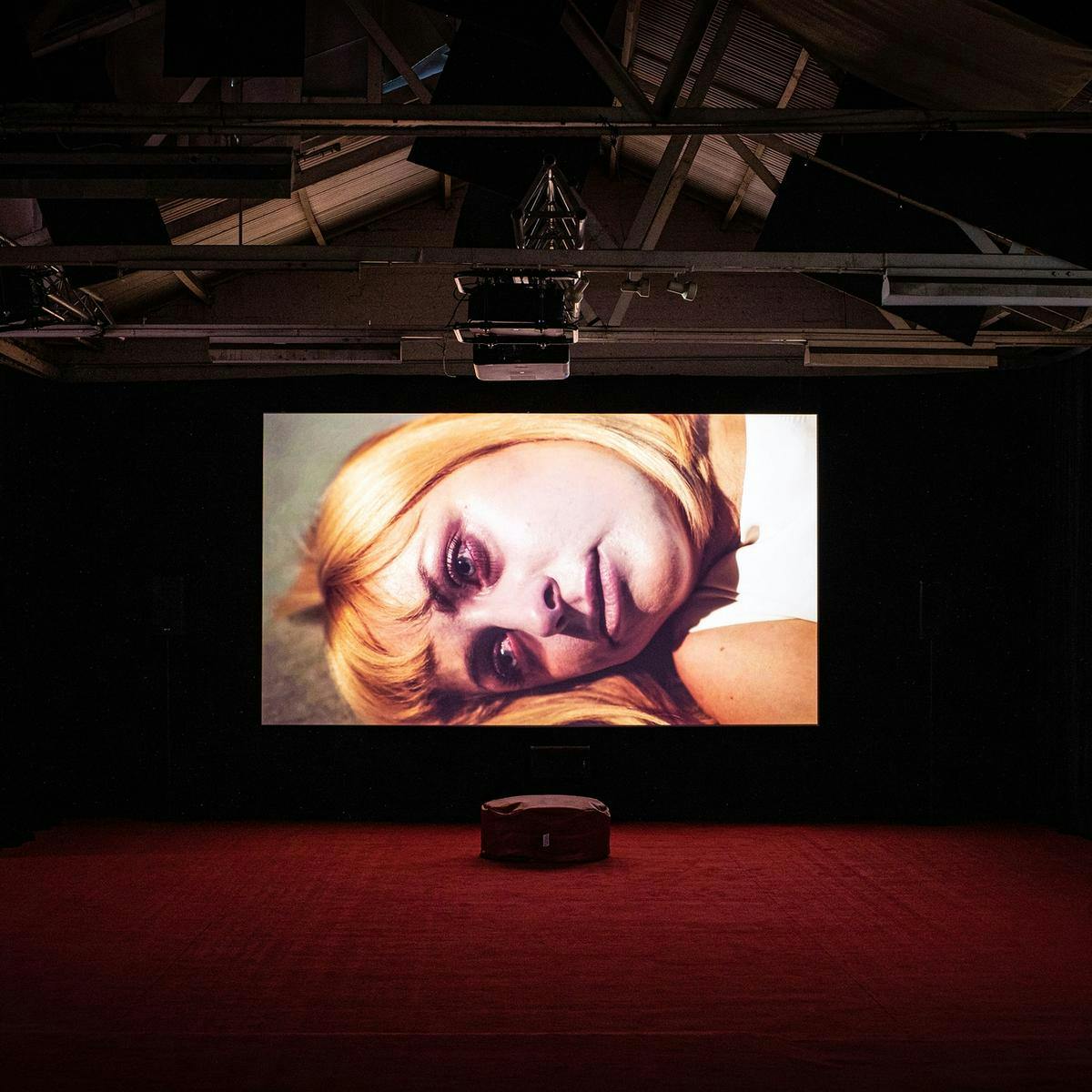 A black walled and red carpeted room with two large screens, on these screens is a closeup of a lying down person's face.