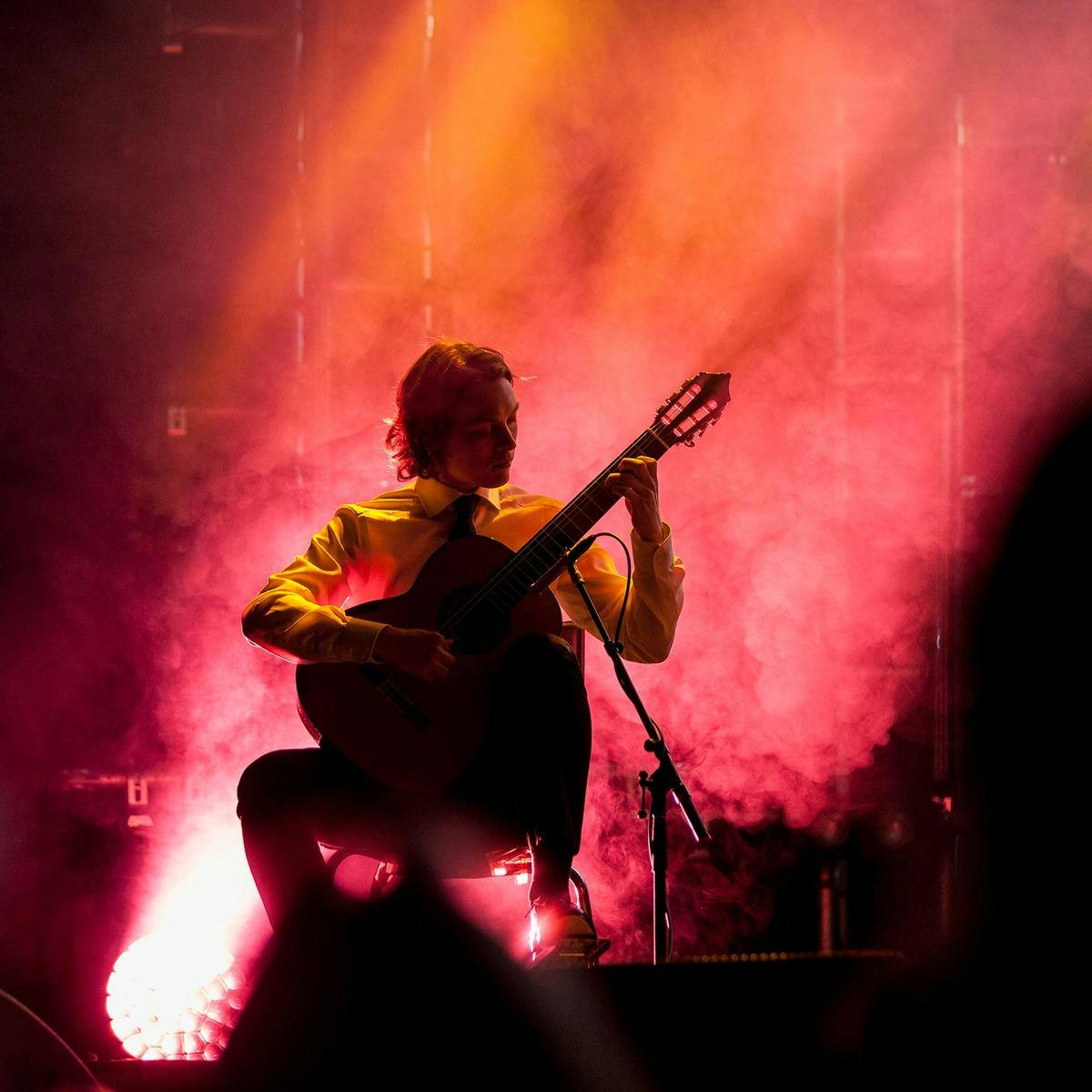 A member of 'True North' performs on stage. Seated, they play an acoustic guitar.