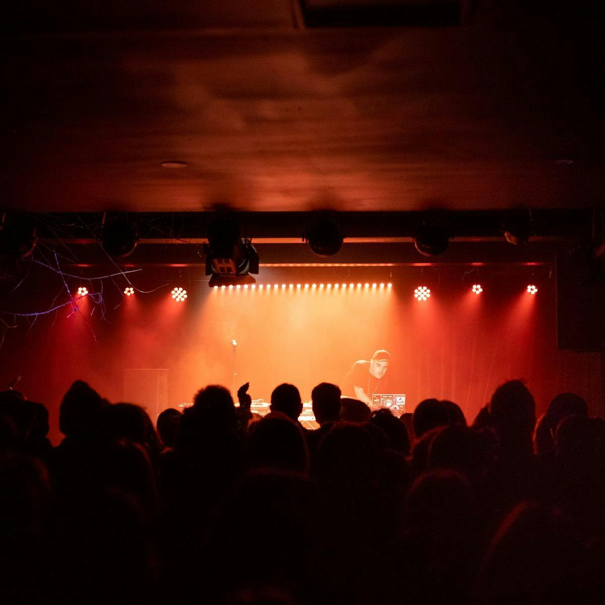 A crowd gathering in a dimly lit room, at the front on stage a DJ attends to their laptop.