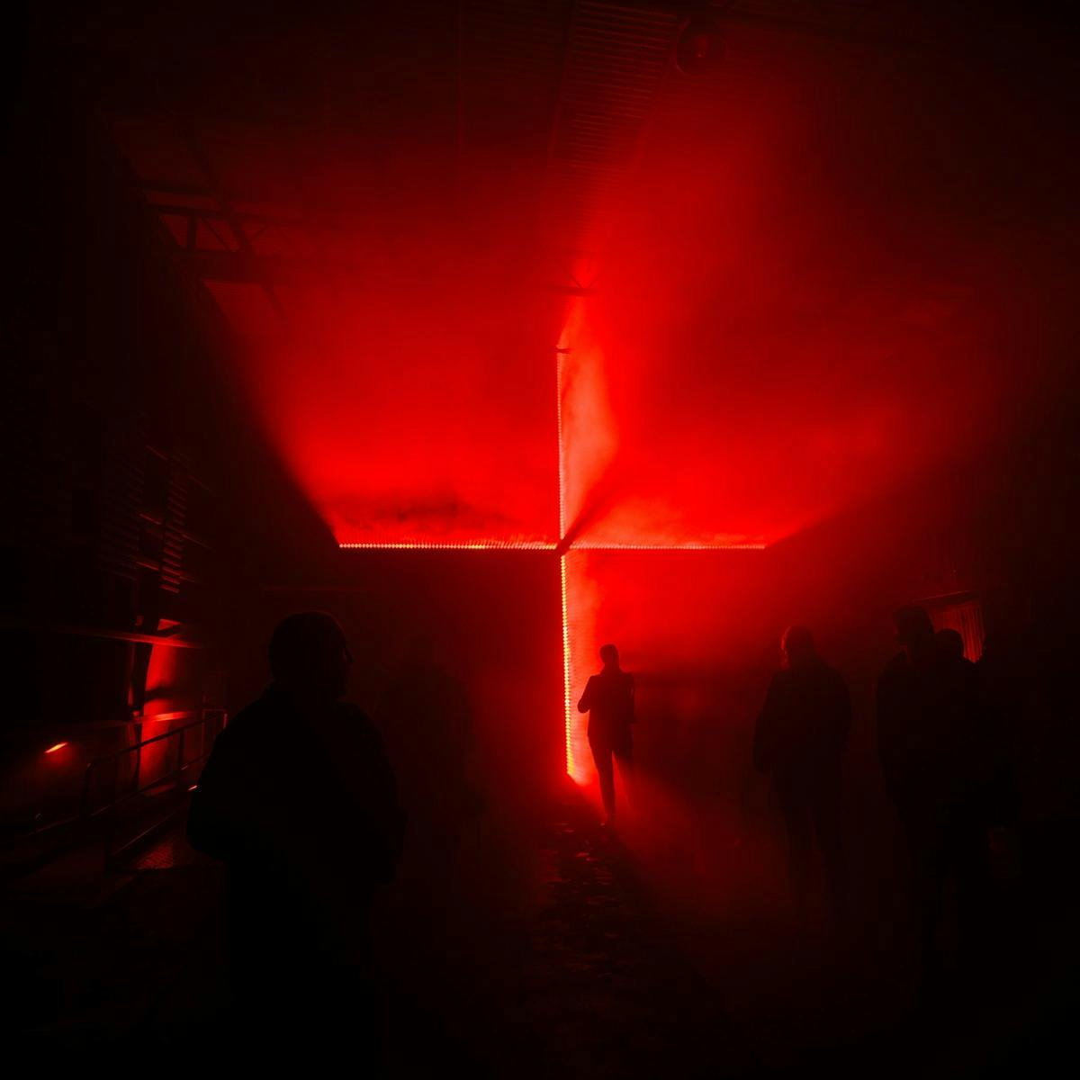 Several people stand in a smoky room, a large equilateral cross made up of lights adorns the back wall.