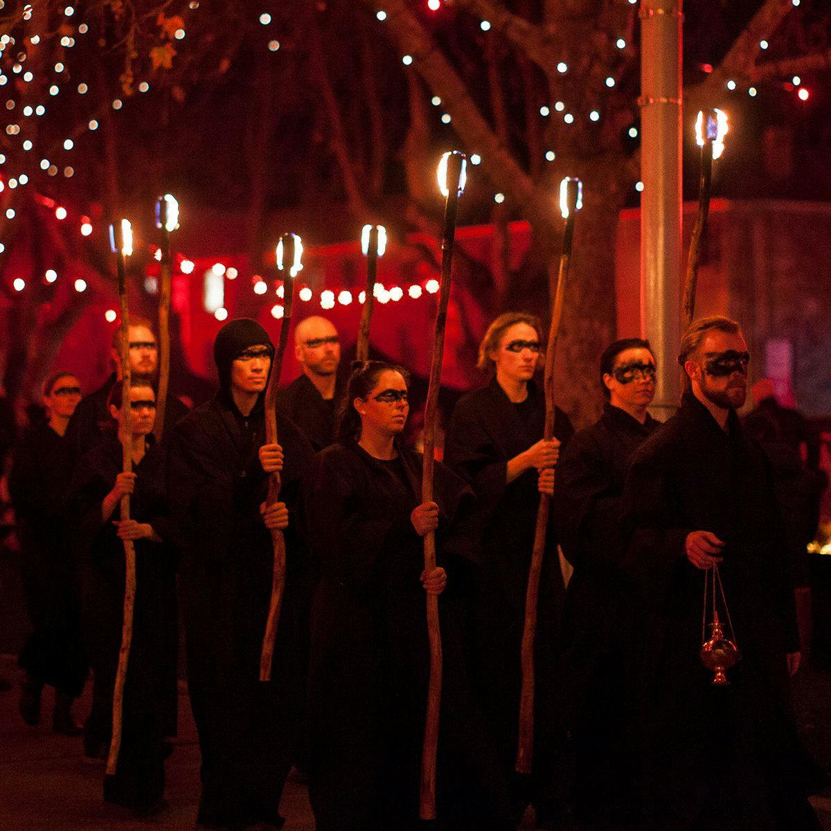 Several people with black face-paint around their eyes carry fire torches. Behind them several trees are lit up with hundred of fairy lights running through the branches.