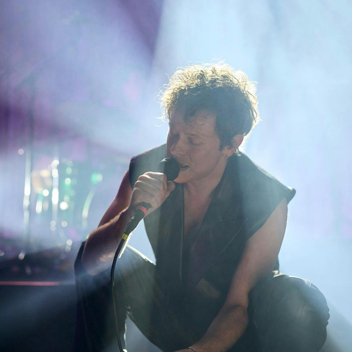 Perfume Genius squats down on stage as they perform into a microphone.