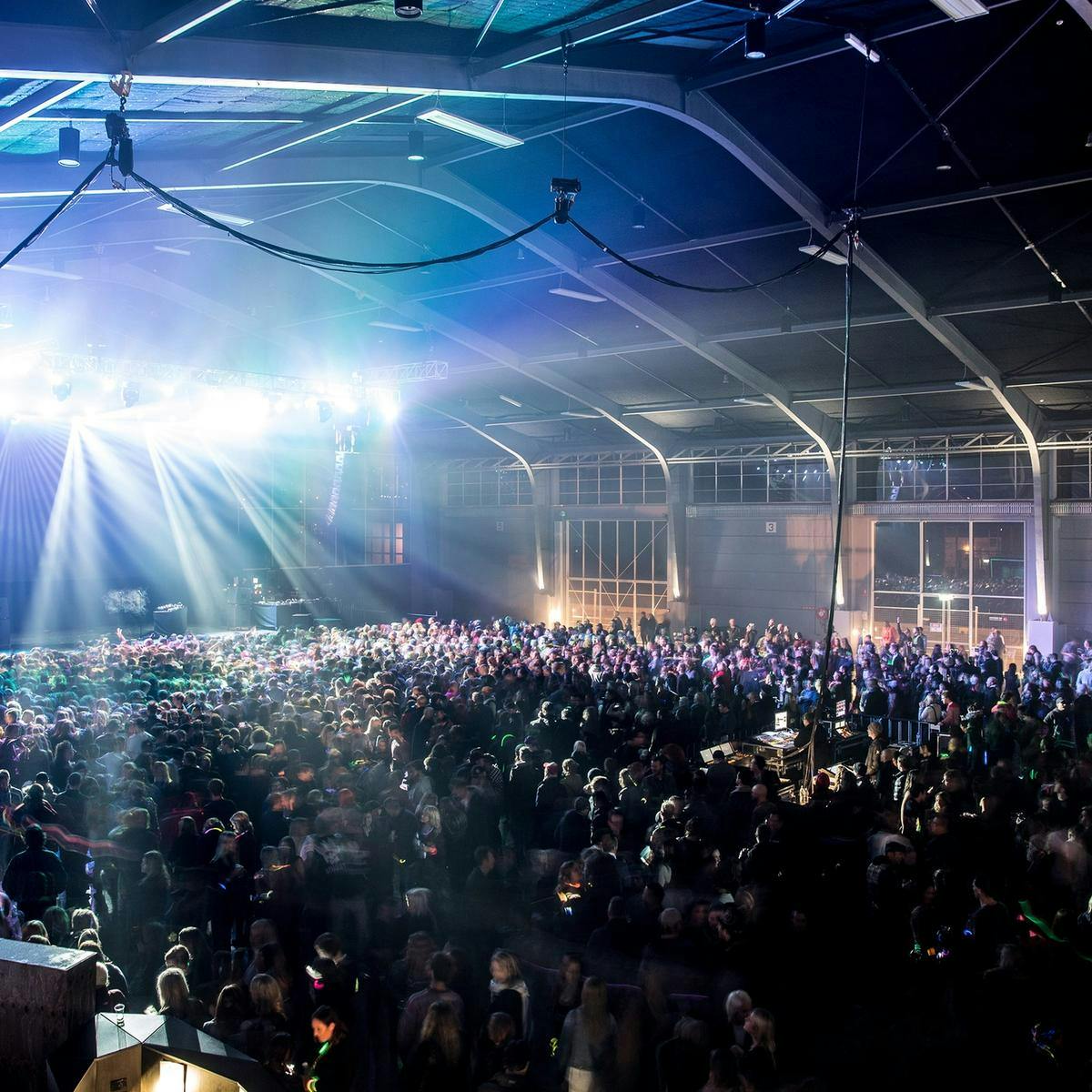 A large crowd of hundreds packs out a warehouse venue. Bright white lights shine outwards from the stage area.