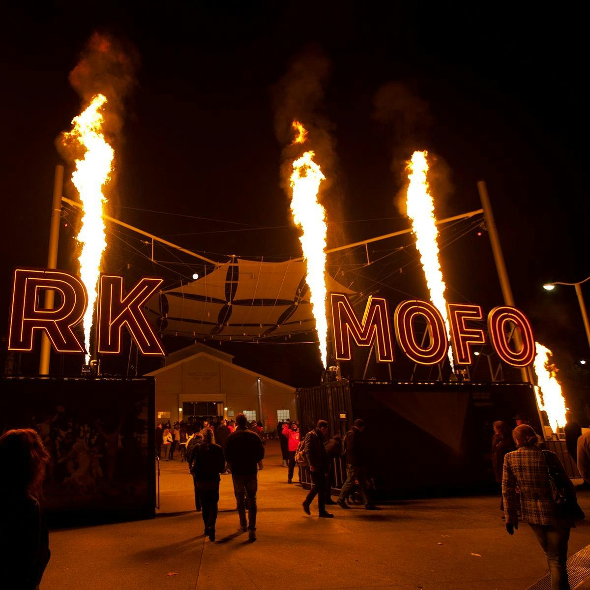 The words 'Dark Mofo' in large neon letters sit on containers, with pillars of fire shooting up behind them.