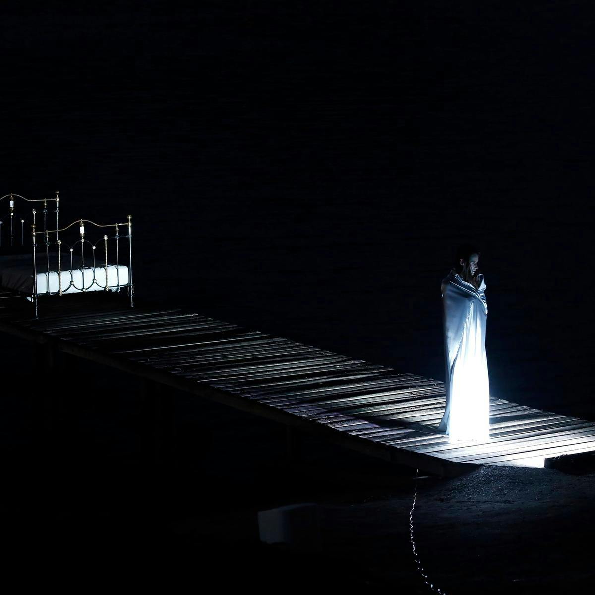 Shrouded by darkness and wrapped in a white shroud, a lone figure stands at the entrance to a wooden pier, at the end of a pier is an ornate metal bed.