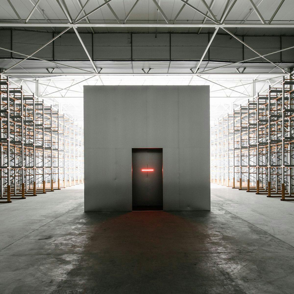 A warehouse-like building with several rows of shelving along the edges. Centered is a white box room, with a closed door.