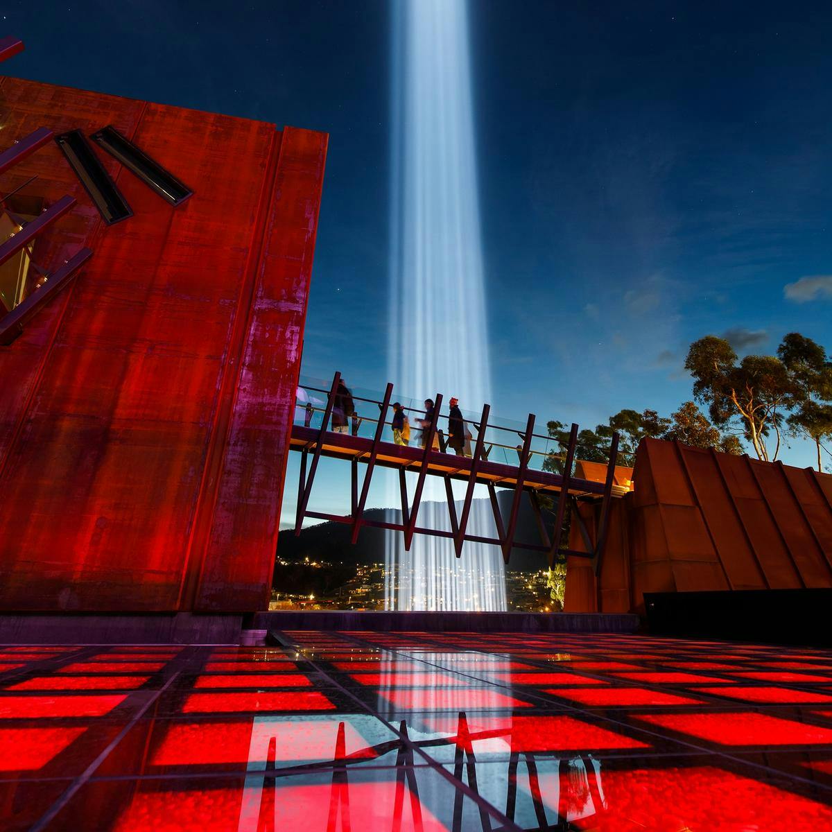 'spectra' beams into the sky at Mona. Several people walk up a pathway leading up to a building.