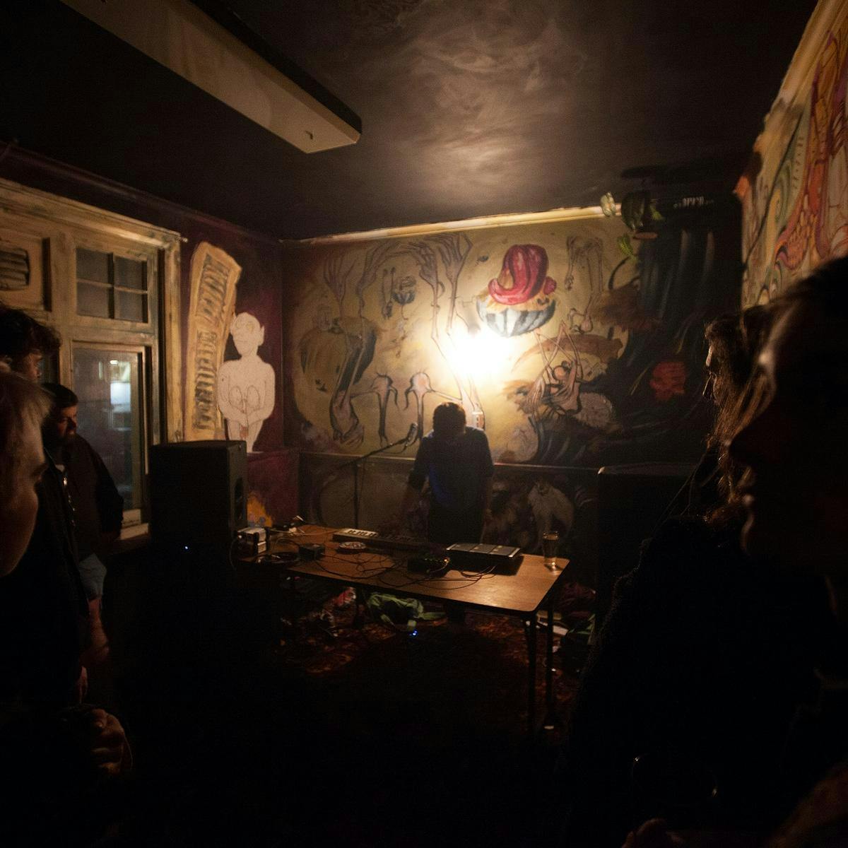 Several people gathered in a grungy, dimly lit room. A DJ performs on a wooden table.