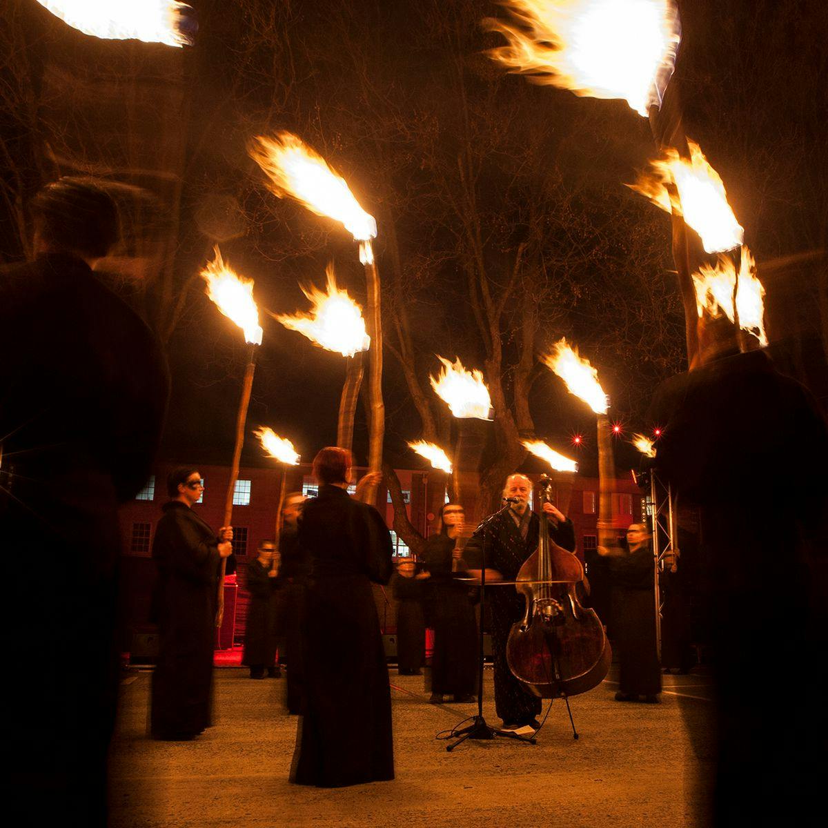 Several figures clad in black robes carry flaming torches, centered is a person playing a cello.