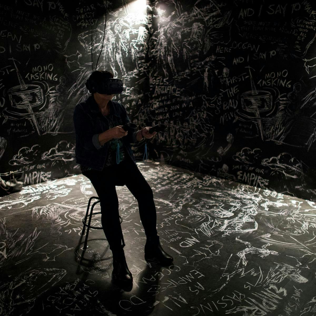 A person sits on a stool wearing a VR headset and holding VR controllers. The black room, is floor to ceiling covered in white chalk drawings and writing.