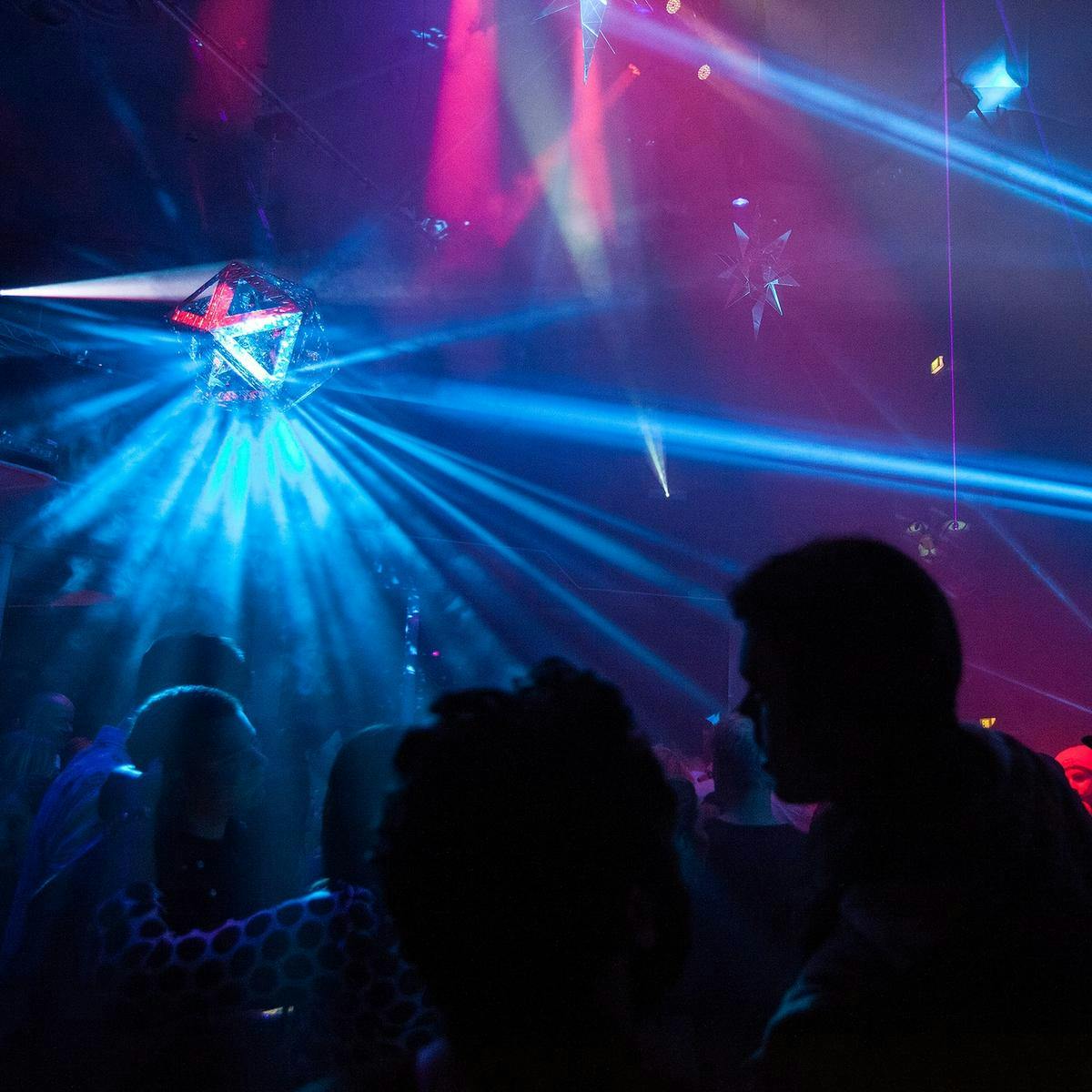 A crowd of people dance and gather beneath a disco-ball like object, which is projecting several beams of bright blue light.