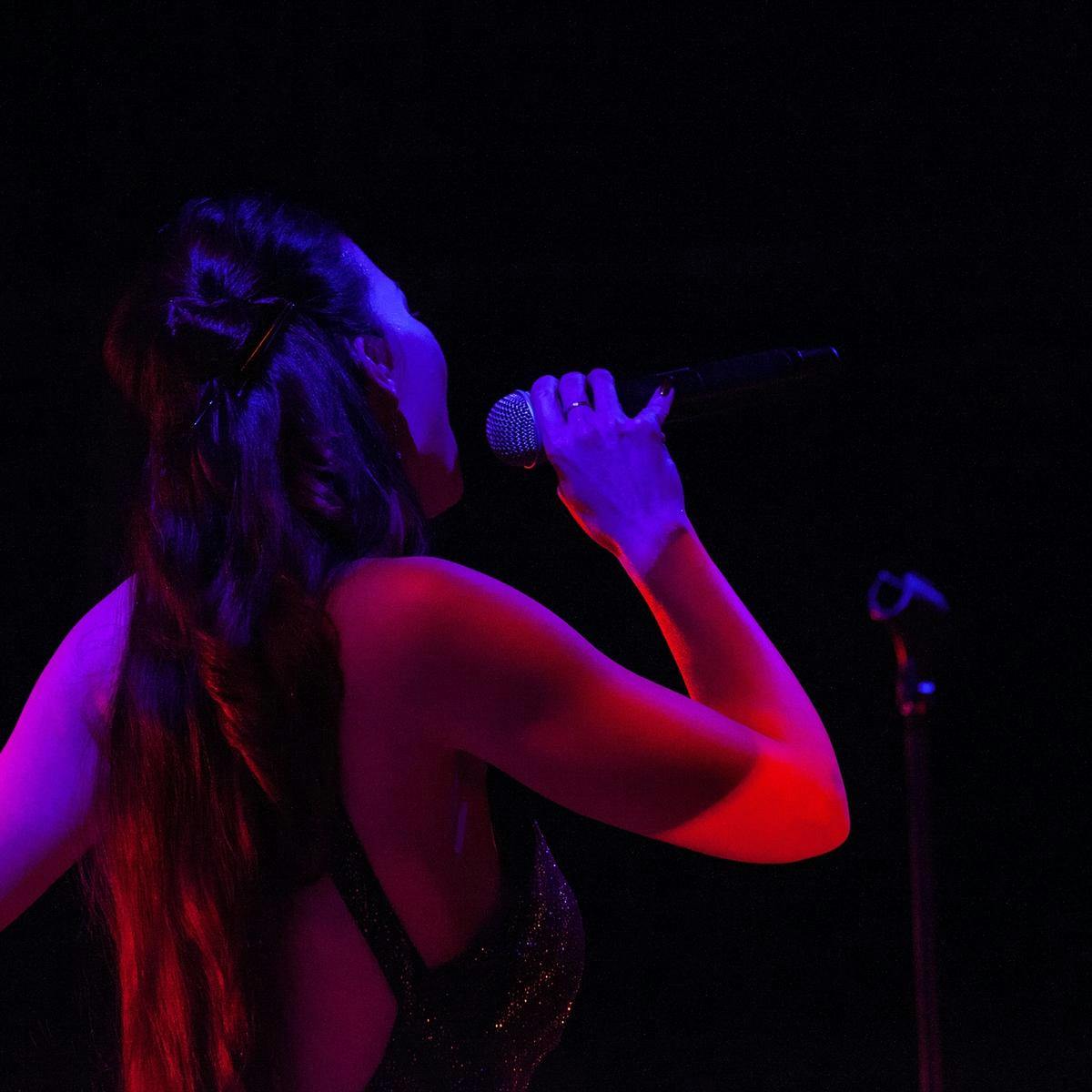 Chrysta Bell performs on-stage singing into a microphone, their body lit up by red and purple lighting.