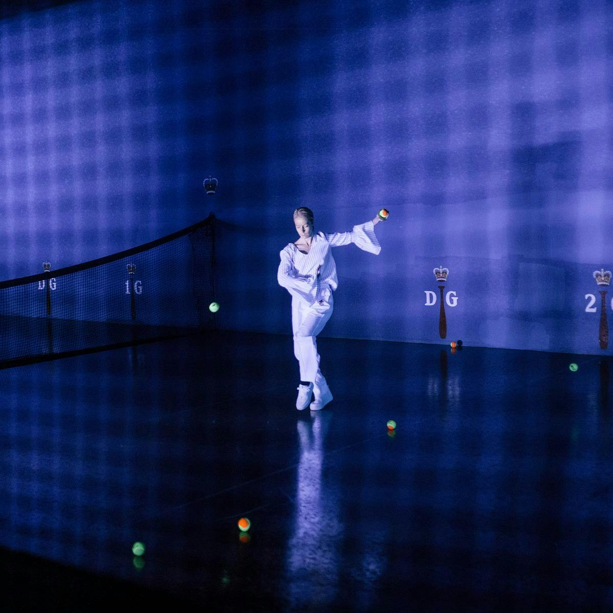 A performer holds a tennis ball, whilst prancing on an indoor court. Several other tennis balls are scattered on the floor.