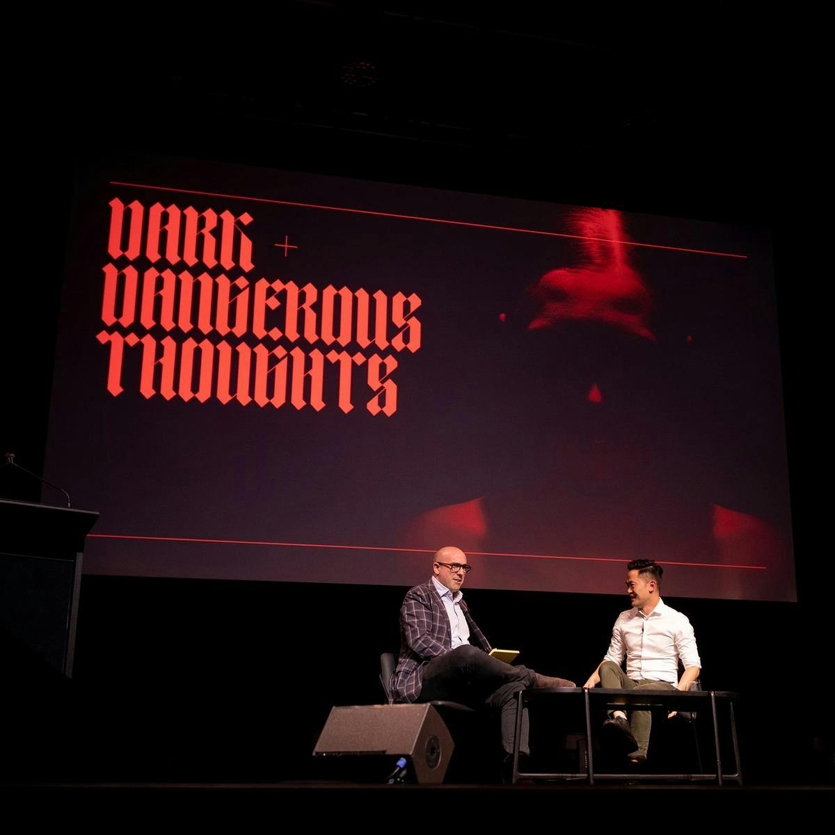 Two people sit on stage in conversation, behind them on a large screen reads 'Dark + Dangerous Thoughts'.