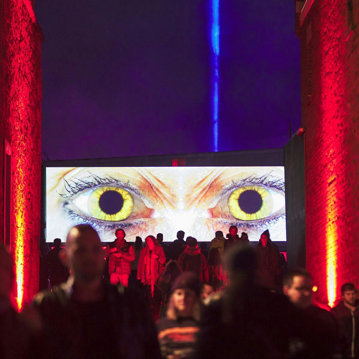 Several people walk through a wide alleyway, above them in the background is a large bright screen, depicting a close up of something's eyes, this is the artwork 'Double Eclipse'.