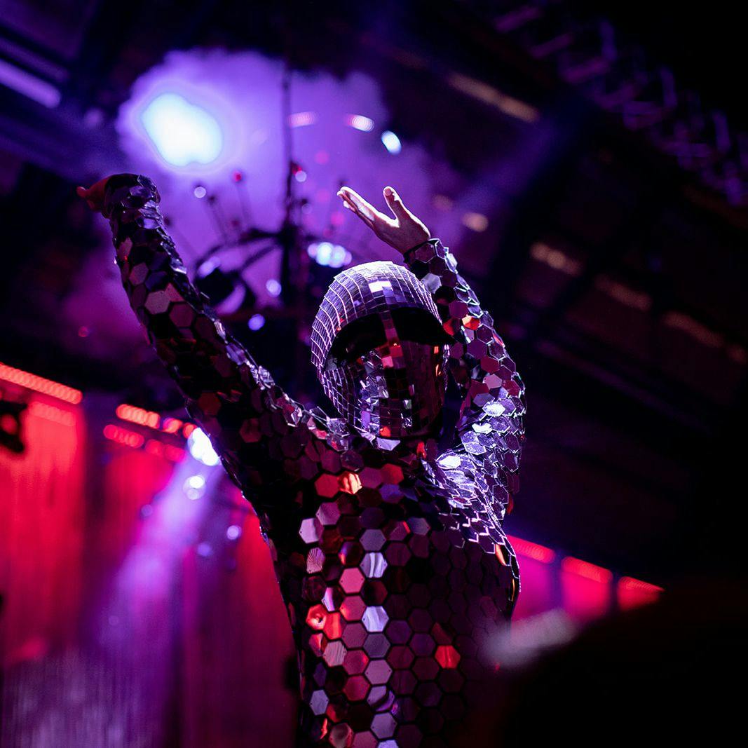 A performer with their hands raised above their head. Their outfit, including mask is made up of hundreds of mirrored shapes.