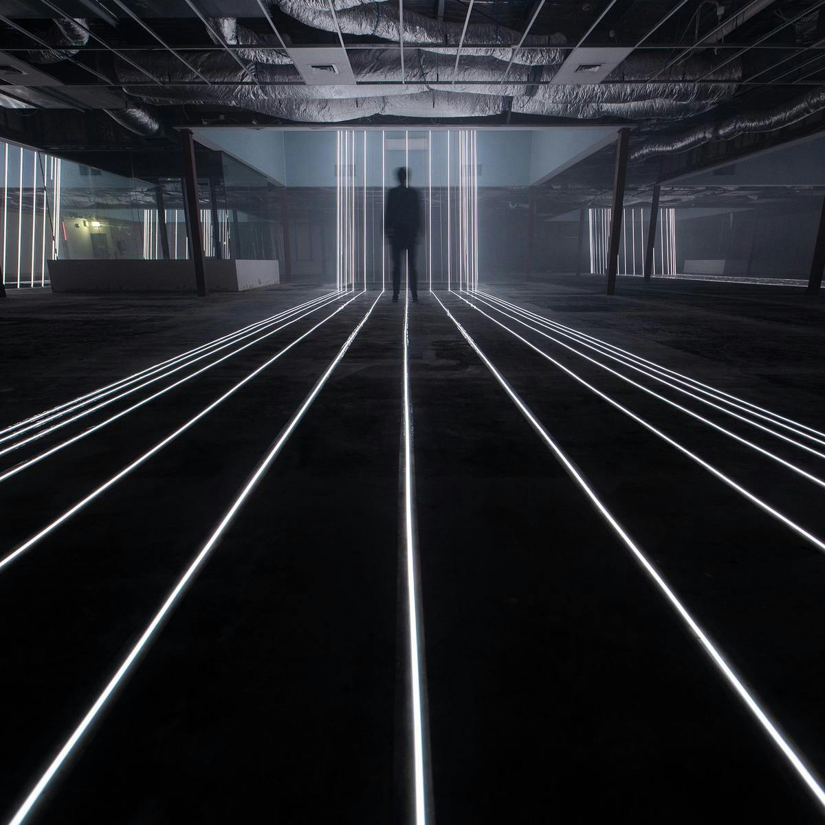 A blurred person stands in the distance, several white strips of light run along the floor towards them, before changing direction towards the ceiling.