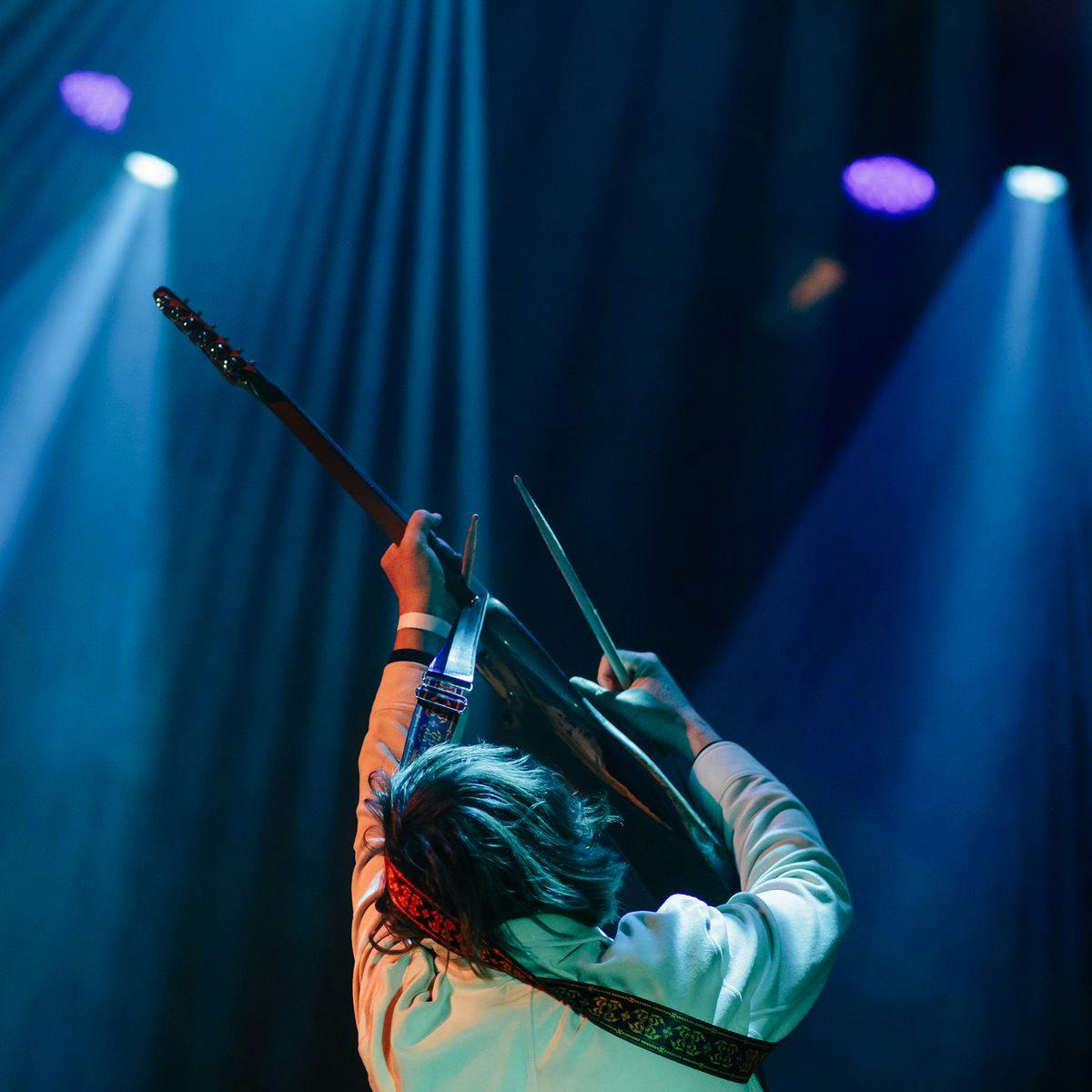 Thurston Moore plays the guitar above their head, whilst holding a drumstick in each hand.