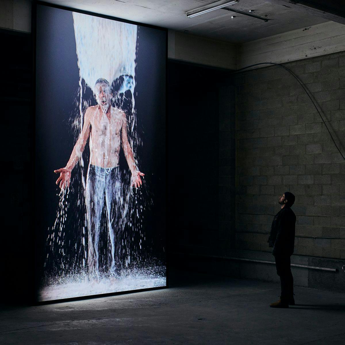 A lone person looks up to a large two-story screen. On the screen footage of a shirt-less man being doused in white liquid plays. This is the artwork 'Inverted Birth'.