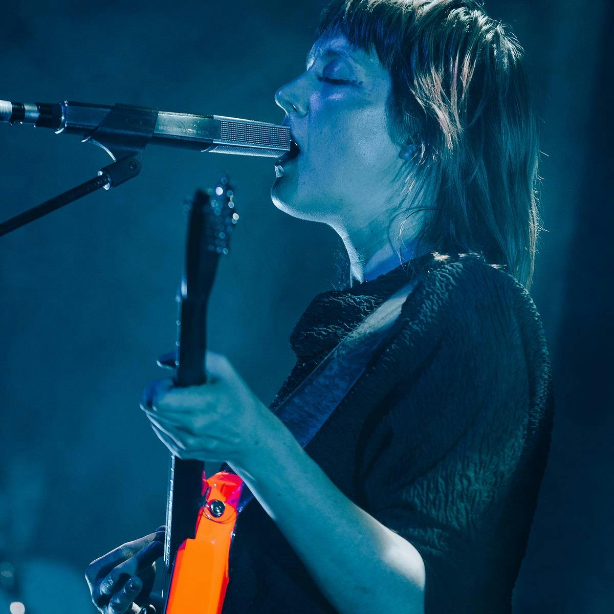 Cate Le Bon performs into a microphone, whilst playing a bright orange guitar.