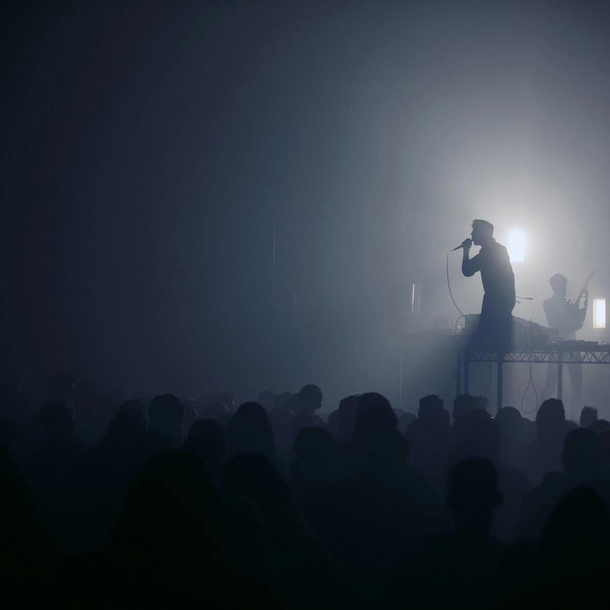 A silhouetted person kneels on a raised table, performing for a large crowd. White light backlights the performer.