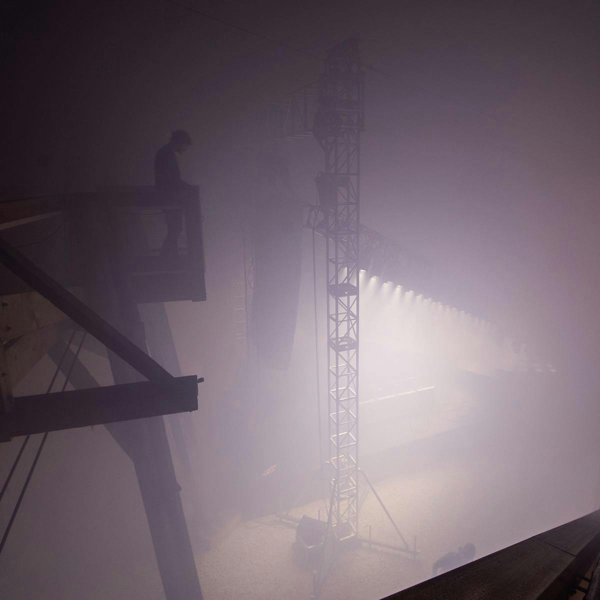 A lone figure stands on a raised walkway, looking down to a mysterious scene of metal scaffolding, white lights and fog.