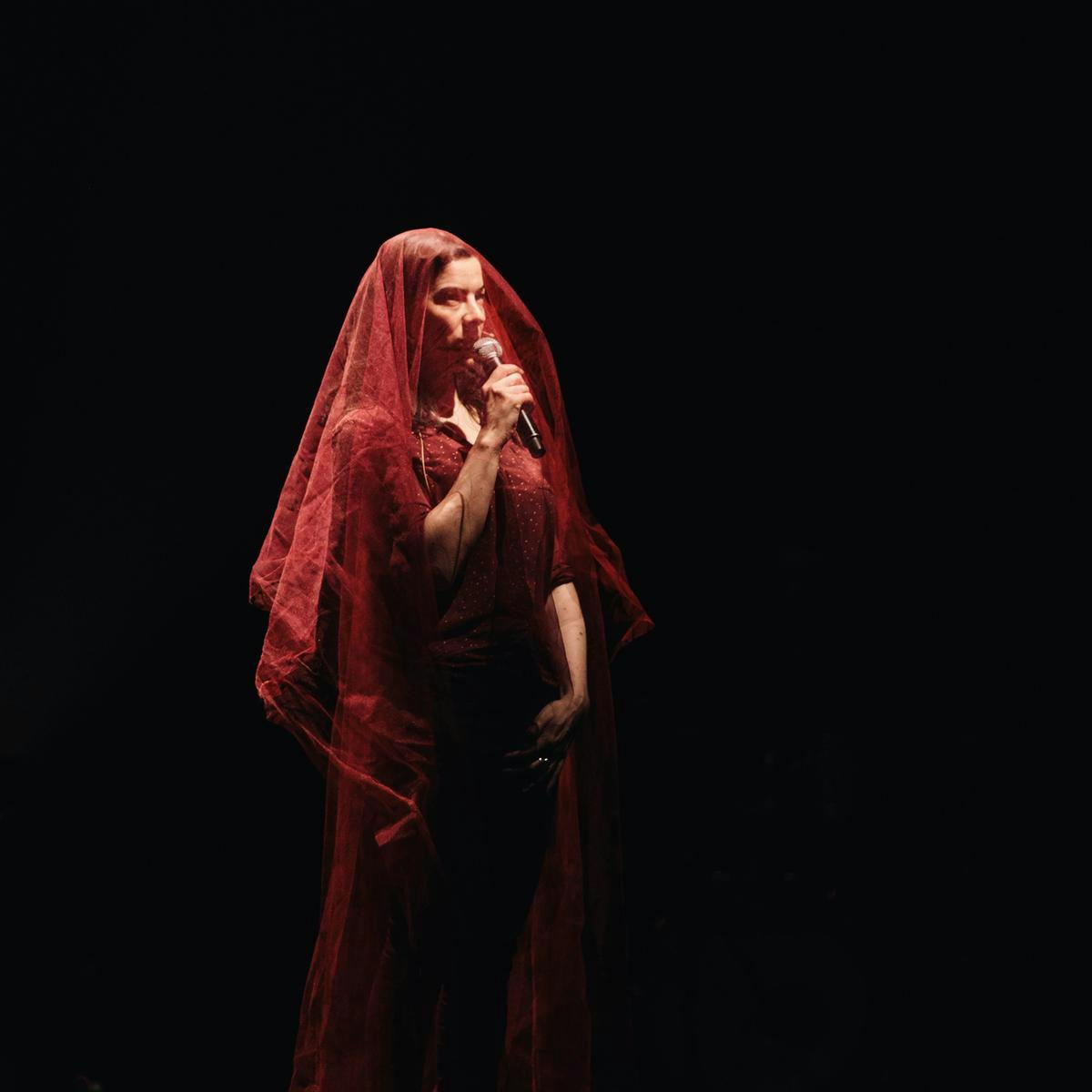 A figure performs into a microphone whilst wearing a flowing red dress and veil.