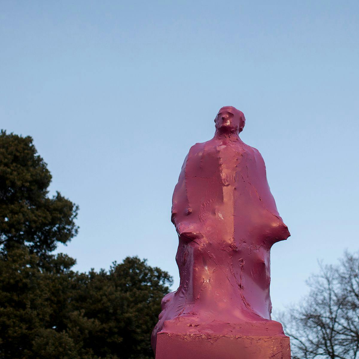 A large statue is shrink-wrapped with pink plastic. Clear blue sky and a few trees are in the background.