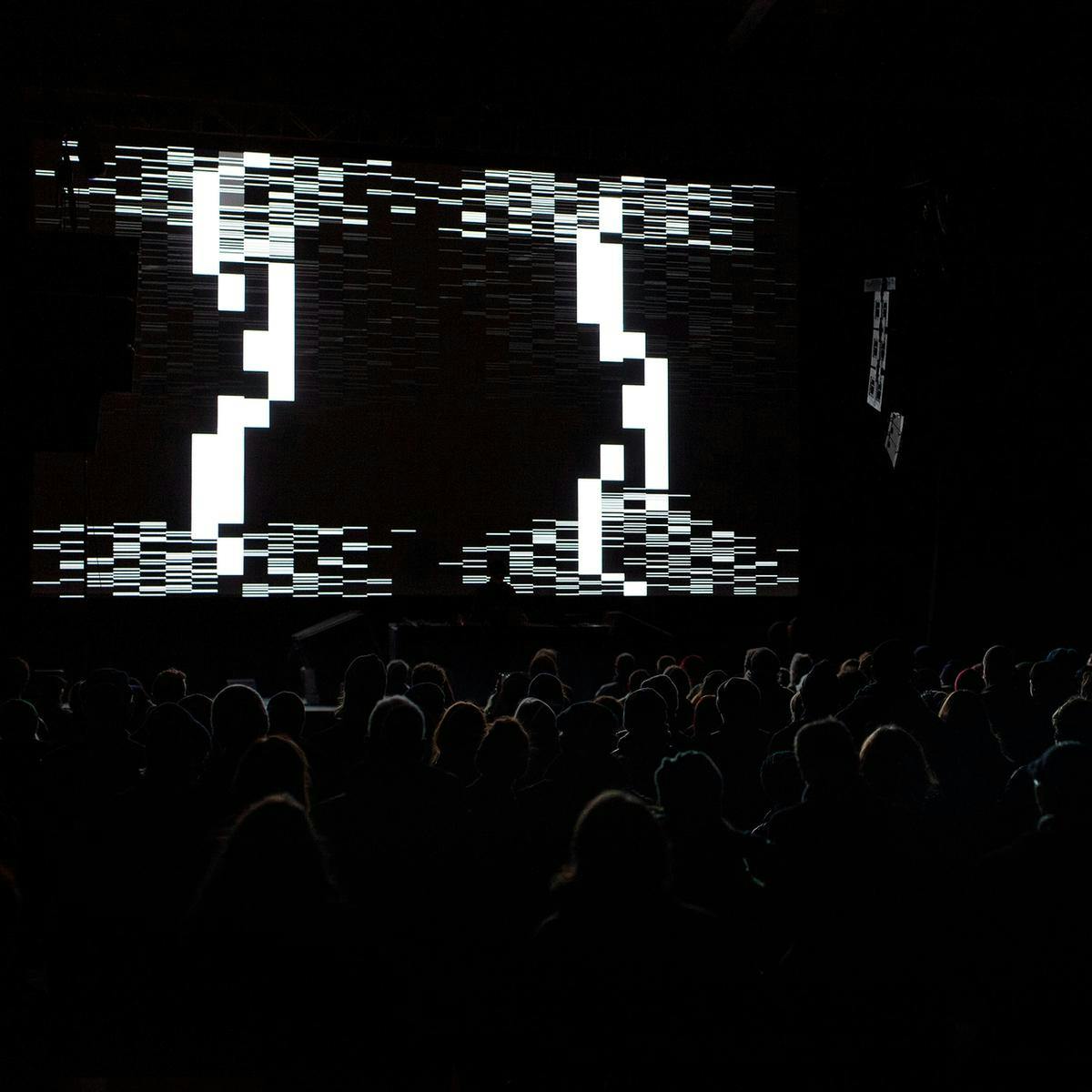 A large crowd face towards a stage, projected behind the stage is an abstract heavily pixelated image.