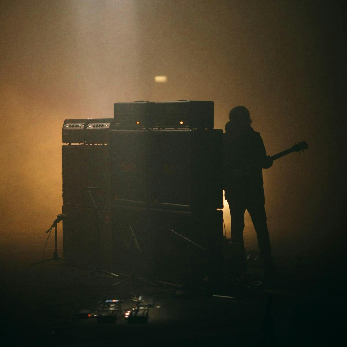 A person holding a guitar stands hidden behind a tall bank of speakers.