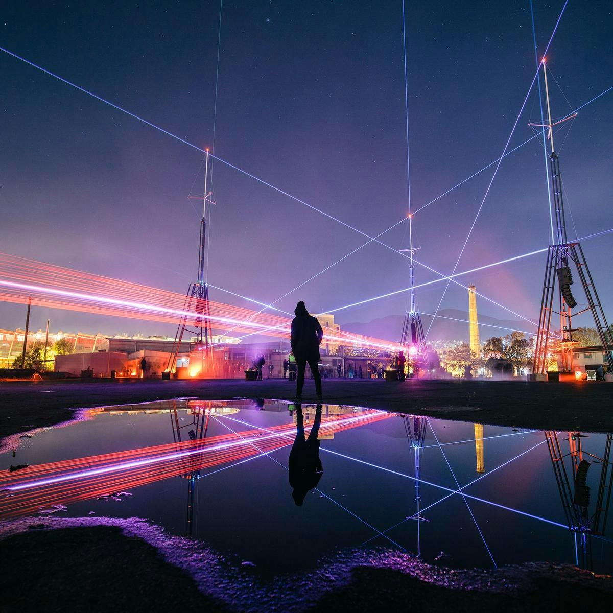 A singular person stands in-front of a large puddle, in the background and reflected in the water is the night's sky and several lasers shooting in many directions.