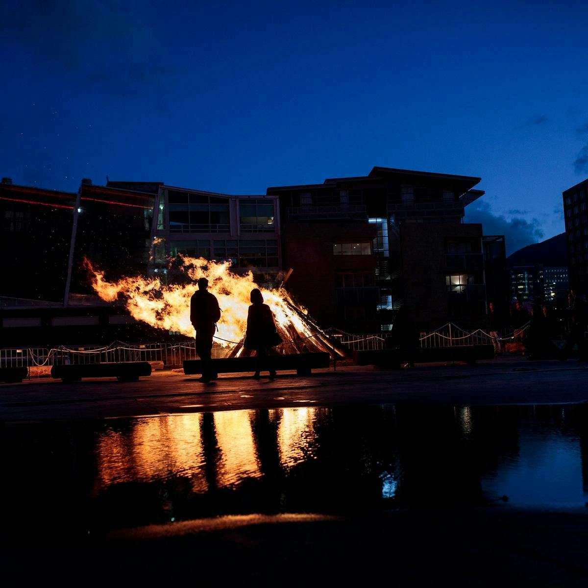 Several people watch a large pyre burning in the night, city buildings are in the background.